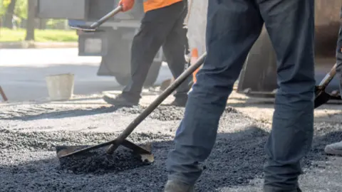 A pair of legs in dark trousers and a large hoe pushing gravel on a road with another person in an orange reflective jacket in the background
