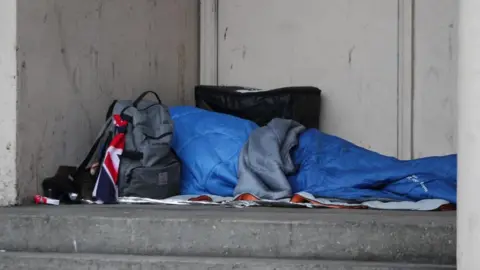 PA Media A homeless person sleeping rough in a doorway. They have a grey rucksack and blue sleeping bag. Their face is not visible.