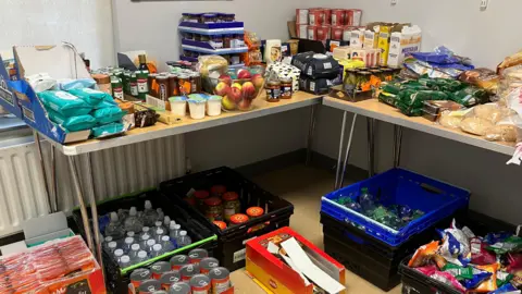 A corner of a room is filled with a colourful collection of cupboard items. In the foreground there are boxes of bottled water and packets of crisps. On the table sits a bowl of apples; loaves of many varieties of bread; cereals; jams ; pots of custard and ready made porridge in pots.