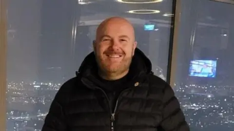 Lee McCormick - a bald man with a beard is smiling at the camera. He is wearing a black puffer jacket and a black tshirt and is stood in front of large glass panels. In the background are lights of nearby buildings and cars.