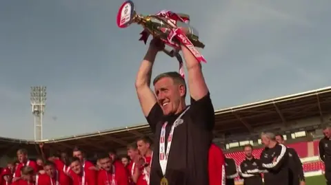 A man wearing a black polo shirt lifts a trophy above his head. He is wearing a medal around his neck. A group of men wearing red shirts stands behind him. They are on the pitch inside a stadium.