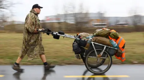 A man wearing a green camouflaged poncho, a baseball cap and boots walks along a wet road, pushing a two-wheeled trolley containing all of his possessions.