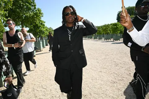 BERTRAND GUAY / AFP / GETTY IMAGES A man dressed in black walks through the Orangery of the Palace of Versailles. He is flanked by an entourage.