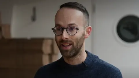 A man with round glasses, short dark hair and a beard looks into the distance. He wears a dark jumper. 