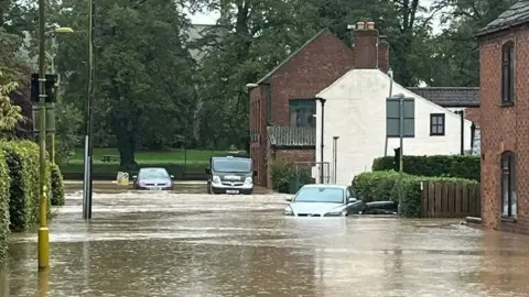 Brown floodwater covers a thoroughfare  successful  Horncastle, Lincolnshire. Two cars and a taxi and partially submerged. To the right, the h2o  laps against red-brick and achromatic  houses to the right. To the left, hedges emergence  supra  the water.