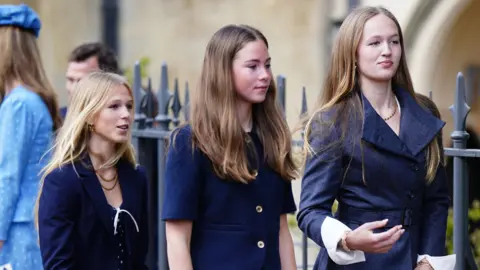PA Three teenage girls wearing formal clothes in dark blue, walking along the street beside an iron railing. 