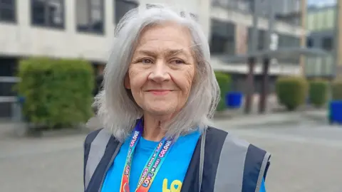 A woman with grey hair is smiling. She is not looking directly at the camera. She is wearing a navy visibility vest and blue t-shirt.