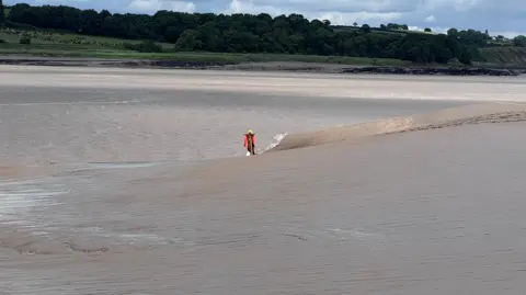 A mannequin wearing hi-vis clothing is nailed to a stake on a river bank. On the other side of the river is lush green farmland and woodland.