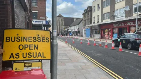 Midland Road in Bedford, showing a sign that says Business Open As Usual, red traffic cones, a black taxi driving along the road, two lanes of traffic and shops down both sides. 
