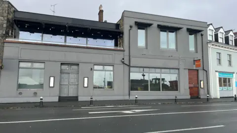 BBC Two grey buildings attached to eachother. the one of the left has a grey door and a balcony and the one on the right has a brown door.