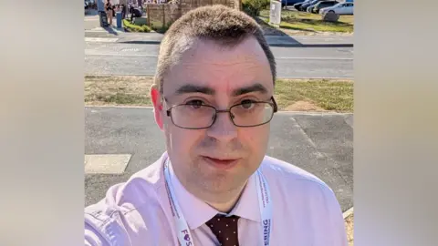 Jamie Baggaley A selfie of a man with brown hair, brown eyes and thick eyebrows. He wears a black shirt, burgundy tie and thin-rimmed black glasses. He has a neutral expression.