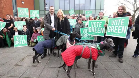 PA Media Four greyhounds on dog leads and wearing jackets stand in front of Green Party leader Zack Polanski, Hannah Spencer and campaigners who are holding signs which read 
