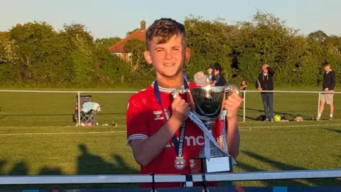 Supplied A teenage boy stands on a football pitch, holding a large silver trophy. He wears a red sports top and a medal around his neck. 