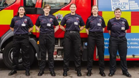 Oxfordshire Fire and Rescue Service Five women in firefighter uniforms stand to attention beside a fire appliance