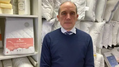 Fraser Smith, who has short, grey hair, is wearing a dark blue jumper over a white shirt with a navy-and-pink striped tie. He is standing in front of a shelf full of cushions and bedding.