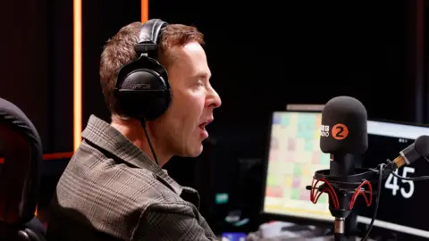 A profile image of Scott Mills wearing headphones while speaking into a microphone with the BBC logo and the number 2 on it, with computer screens and the studio in the background, in early 2026.