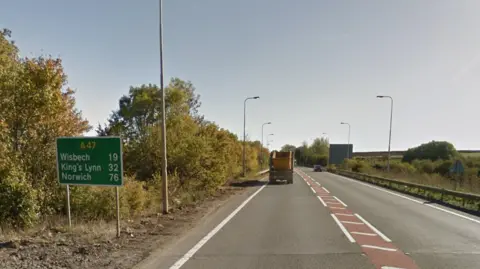 A section of straight A47 single carriageway road, with white road markings and a green highways sign to the left which reads A47 in yellow, and, in white lettering, Wisbech 19; King's Lynn 32; Norwich 76. A truck with a yellow skip on the back can be seen driving off into the distance, and foliage either side of road. 