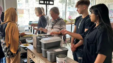 Aishah Ahmed is directing a team of volunteers, who are stood behind a makeshift counter, serving free plates of chicken korma curry and rice to a group of people. Aishah is stood to the right of the photo. She has black hair and wearing a black shirt. On the counter are several large containers holding the curry and a small white metal tin with 'donations' written on it. 