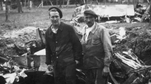 IWM Duxford A black and white photo taken in 1944 of Lt Lonnie Moseley and Lucien Lestang in front of a pile of debris that was Lonnie's US fighter plane. Lonnie is on the left and is wearing a heavy jacket over trousers, with a beret on his head and Lucien is on the right and wearing a cap and a battered heavy cotton work jacket over trousers. 