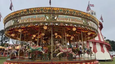 A brightly coloured vintage carousel decorated with lights and painted horses is set up on grass at a fairground, with “Hayward Brothers” written across the top. Union Jack flags fly above the ride, and a large striped tent stands in the background, adding to the traditional fair atmosphere.