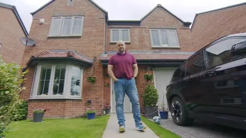 Kevin stands on the footpath leading to the front door of his home. It is a two-storey brick property with a bay window and built-in garage on the ground floor. There is a black SUV car parked on the drive and there are plant pots and hanging baskets outside the home. Kevin is wearing and burgundy polo shirt, blue jeans and trainers. He does not look happy 