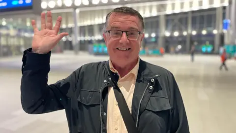 BBC Kieran Doherty stands in Grand Central Station, smiling and waving at the camera. He is wearing glasses, a cream shirt and a black jacket. The background of the photo is blurred. 