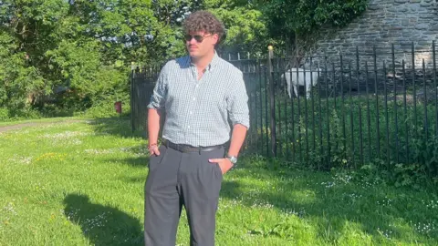 Caleb Tutt Caleb standing on a green lawn. He has sunglasses on and has curly dark hair. He has a green, white and purple checked shirt with black trousers. Behind him is a black metal fence with a white goat.