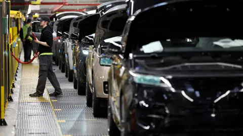 Reuters A worker on a car production line. A line of vehicles all have their bonnets up 