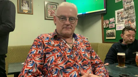 BBC/Lucy Ashton A man with white hair swept back is wearing glasses, stud earrings and a bright red, white and blue Union Jack style shirt with a swirling pattern. He is sitting at a table in a social club with a large television screen in the background