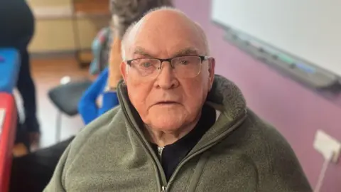 Image of Sean Kerr, at a meeting of the group. He is looking at the camera and wearing glasses and a green zip-up jumper. Some scattered chairs can be seen in the background.