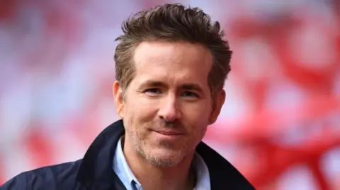 Ryan Reynolds looking directly at the camera, smiling. He has short light brown hair and facial hair. He is wearing a dark navy jacket and a shirt. 