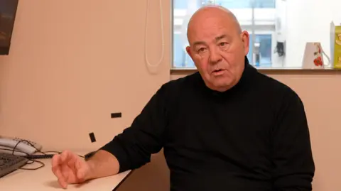 A bald man in a black polo neck sits at a desk and looks at the camera off-centre. A window can be seem behind him and a cup upside down on the window sill.