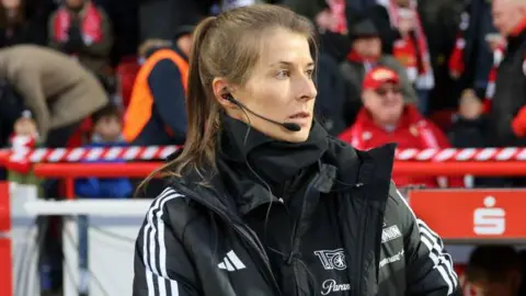 Marie-Louise Eta, wearing a black coat and top with a black microphone and headset in her right ear, on the touchline with fans in red and dark clothing in the background