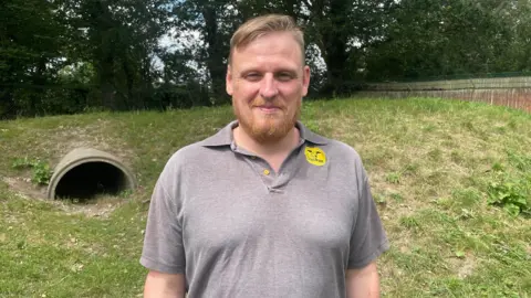 A man with short blond hair and a ginger beard stands on grass, smiling at the camera. He wears a grey polo shirt with a yellow Dogs Trust logo. Behind him are a large concrete pipe, a grassy mound and trees.
