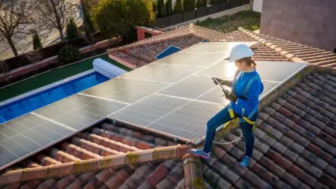 Getty Images A woman works on the maintenance of a solar photovoltaic panel installed on domestic home rooftop. She is standing on the roof and looking at a tablet and it is sunset.