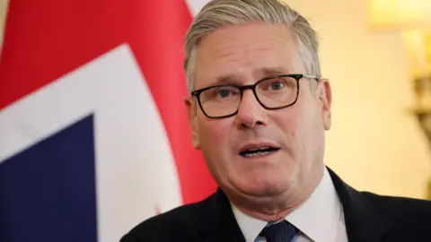 EPA Sir Keir Starmer speaking in front of a union jack flag. It is a head and shoulders shot - he is wearing a white shirt, dark black suit and a dark tie.