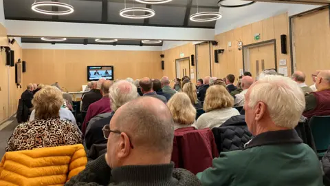 Shariqua AHmed/BBC Members of the public sitting down inside a room with wooden panelling, with their backs facing the camera. They are all looking at a TV screen that is televising a meeting. 