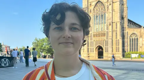 Zoe Dennis/BBC A woman with short wavy brown hair looks at the camera in a head and shoulders image. She is wearing a white tshirt and an orange and cream striped jacket. 