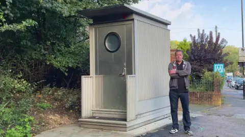 Brett Kemp, a man in his 50s, standing next to a tall grey shed that houses a toilet, erected for the convenience of bus drivers. His arms are folded and he looks cross.