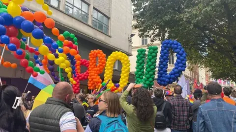 A rainbow collection of balloons spell out "proud" above the heads of a crowd of people on a street.