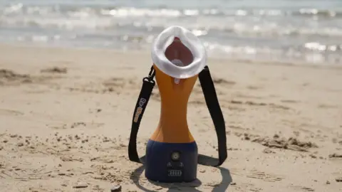 The image shows the device sat on the beach. The sea is in vision behind it. The device has a blue base and the rest is orange. It has a clear mouth and nose piece. It has a black strap on it which says 'Re-Warm'.
