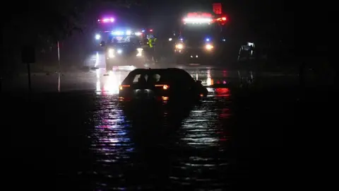 A car is partially submerged in water. It's nighttime and emergency vehicles are in the background with flashing lights.