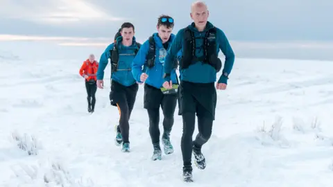 Jamie Rutherford  / Montane Winter Spine Race Three men in blue tops and black leggings running along a snow covered track, with a man in red further back