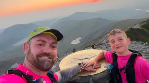 Daniel Thomas Daniel Thomas and Albie-Junior standing at the peak of Yr Wyddfa, also known as Snowdon. An organe and pink sunset can be seen in the horizon. They are both smiling and looking at the camera.