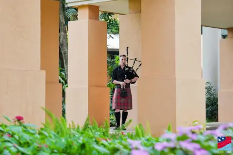 Matt Kidd Matt Kidd plays the bagpipes while wearing a kilt. He is standing outside, in the grounds of a building.