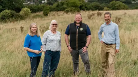Martin Phelps Members of the society and Moreton Cullimore, smiling, whilst standing in an overgrown field with woodland in the background