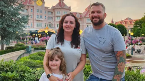 Angharad Dennis Angharad Dennis and her husband Joe are wearing gray T-shirts with the Mickey Mouse logo on them. They stand smiling with their daughter Kari in the Disneyland park.