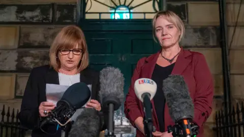 PA Media Two women stand by a podium, one reads off a sheet of paper. The other - Sandie Peggie - looks at the camera. She wears a dark red blazer and has her blonde hair tied back