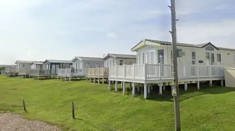 A row of modern static caravans raised on stilts along a grassy slope, each with a white‑railed deck, a gravel path and rope fence in the foreground, and a utility pole standing nearby.