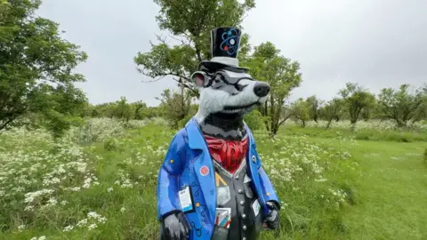 A painted sculpture of Badger from The Wind in the Willows. He is wearing a top hat with an atom painted onto it, a blue blazer, a black waistcoat with various stationery items stuffed into the pockets and a red cravat.